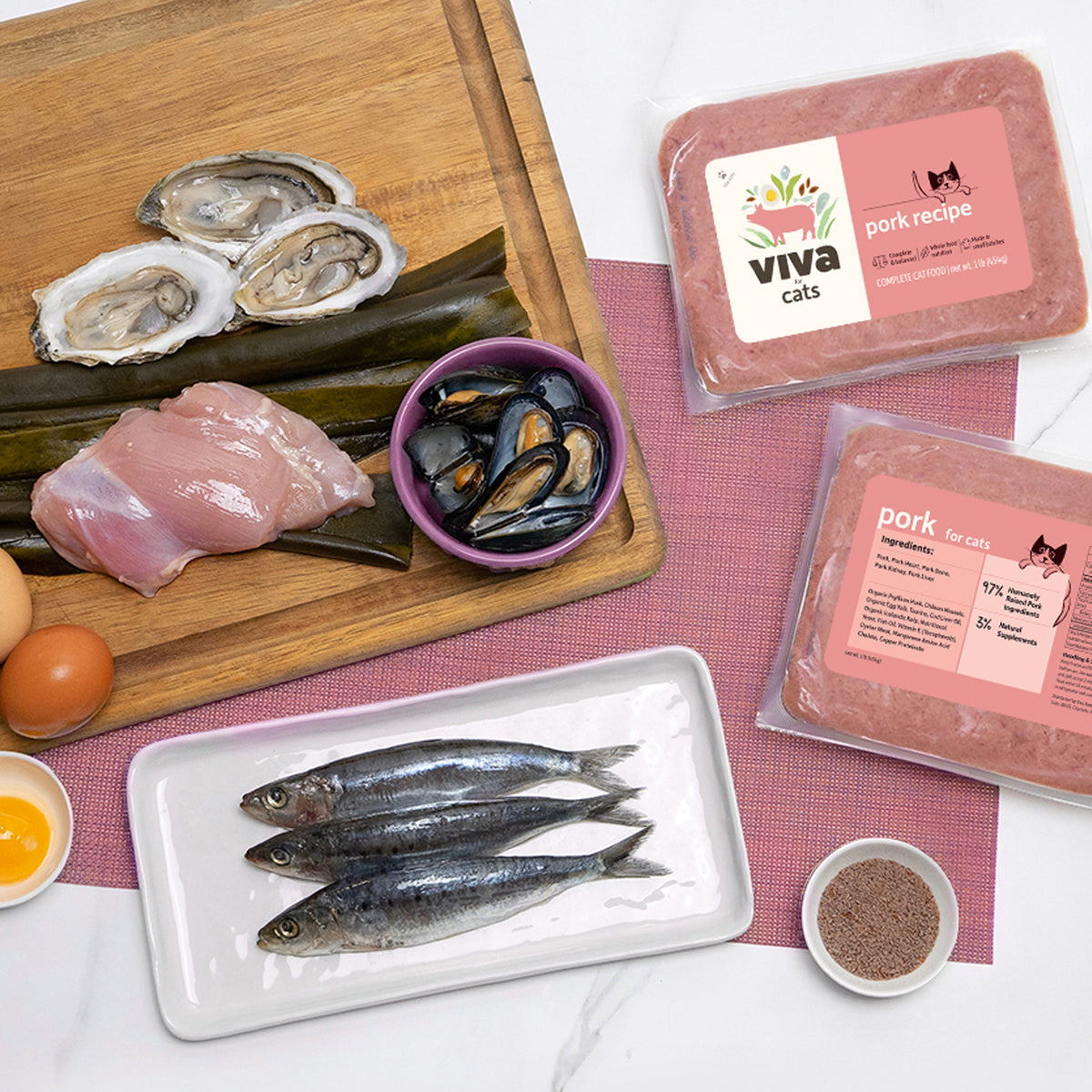 Raw cat food ingredients including pork, sardines, oysters, mussels, and eggs next to Viva for Cats pork recipe packs