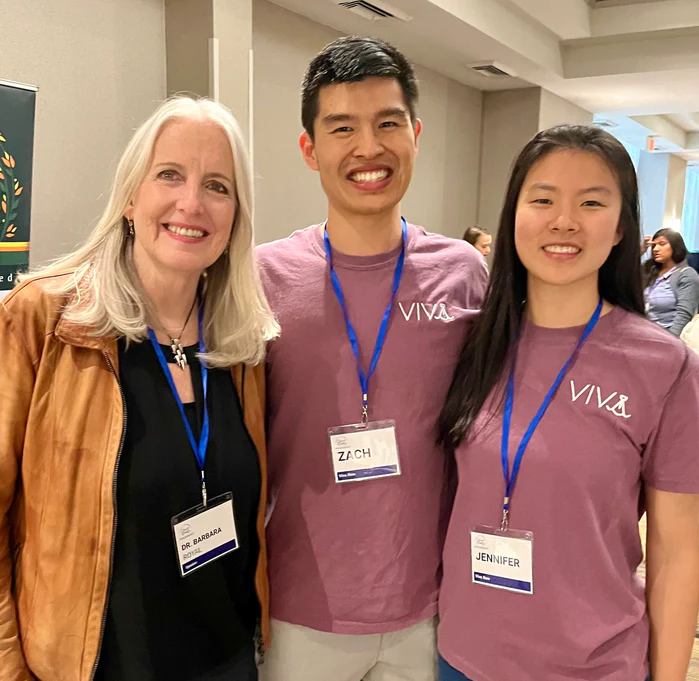 Three people smiling at an event; two wear matching VIVA shirts and name badges, one wears a brown jacket.